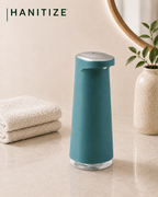 Teal Hanitize automatic soap dispenser on a modern bathroom countertop beside folded towels and a decorative plant.