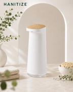 White Hanitize automatic soap dispenser with a wood-effect top on a bright bathroom countertop beside towels and greenery.