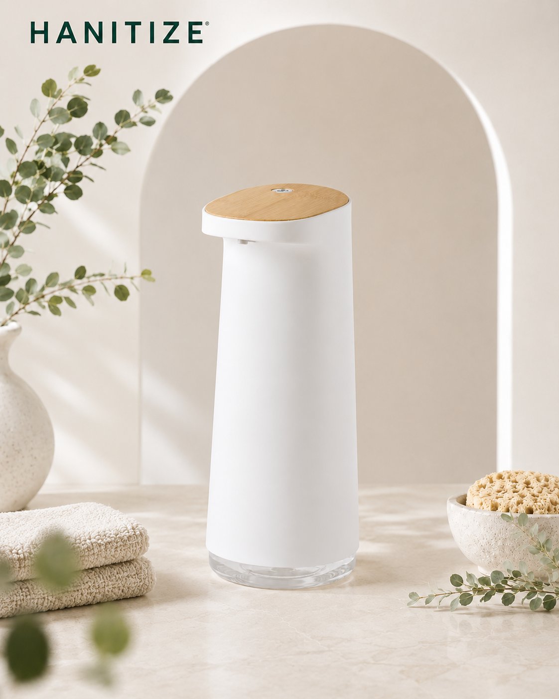 White Hanitize automatic soap dispenser with a wood-effect top on a bright bathroom countertop beside towels and greenery.